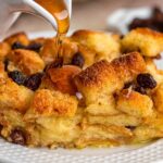 Caramel sauce being poured over bread pudding with raisins on plate.