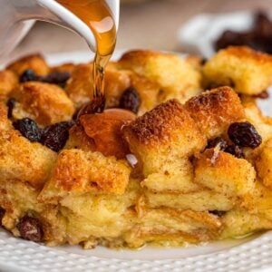 Caramel sauce being poured over bread pudding with raisins on plate.