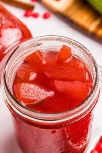 Jar of red cinnamon pickles sitting on a towel with a cinnamon stick.