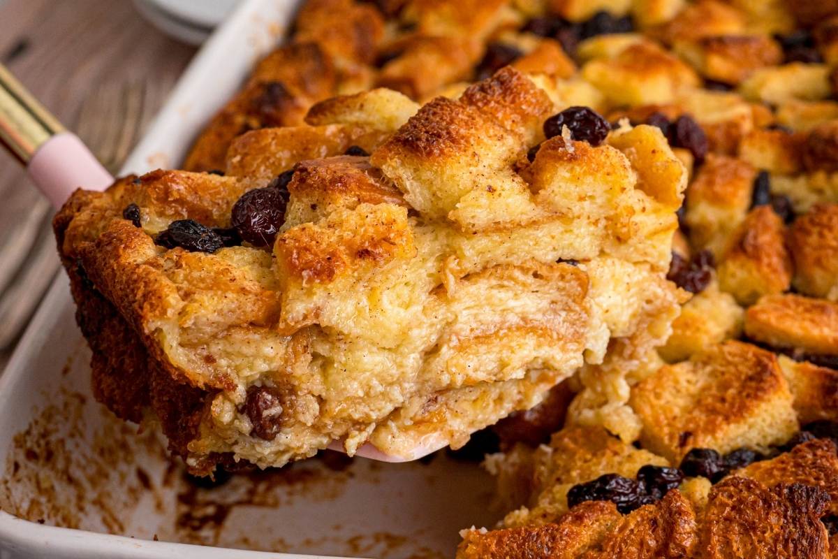 Serving spoon lifting a piece of golden bread pudding with raisins.