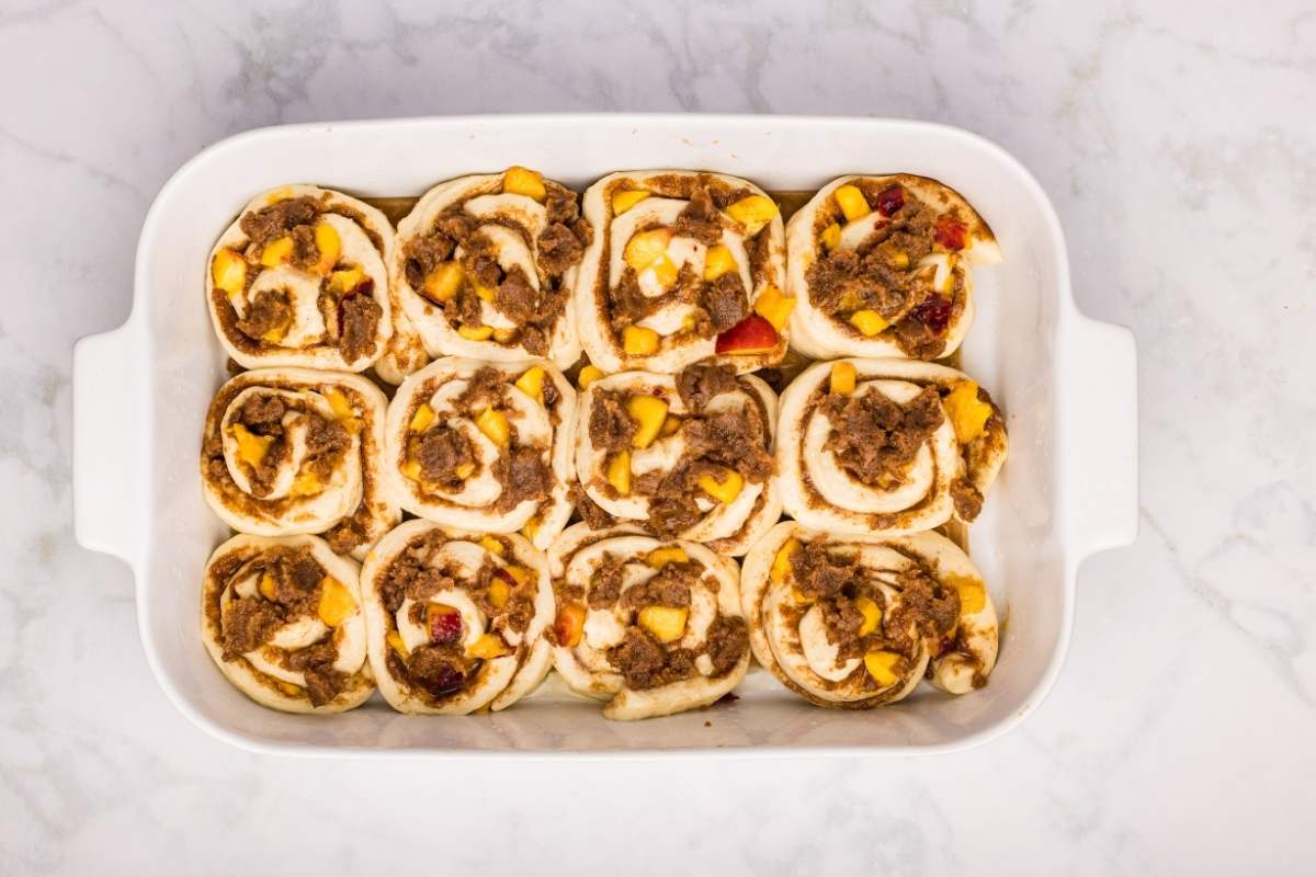 Unbaked cinnamon rolls with peaches and crumble in a white dish.