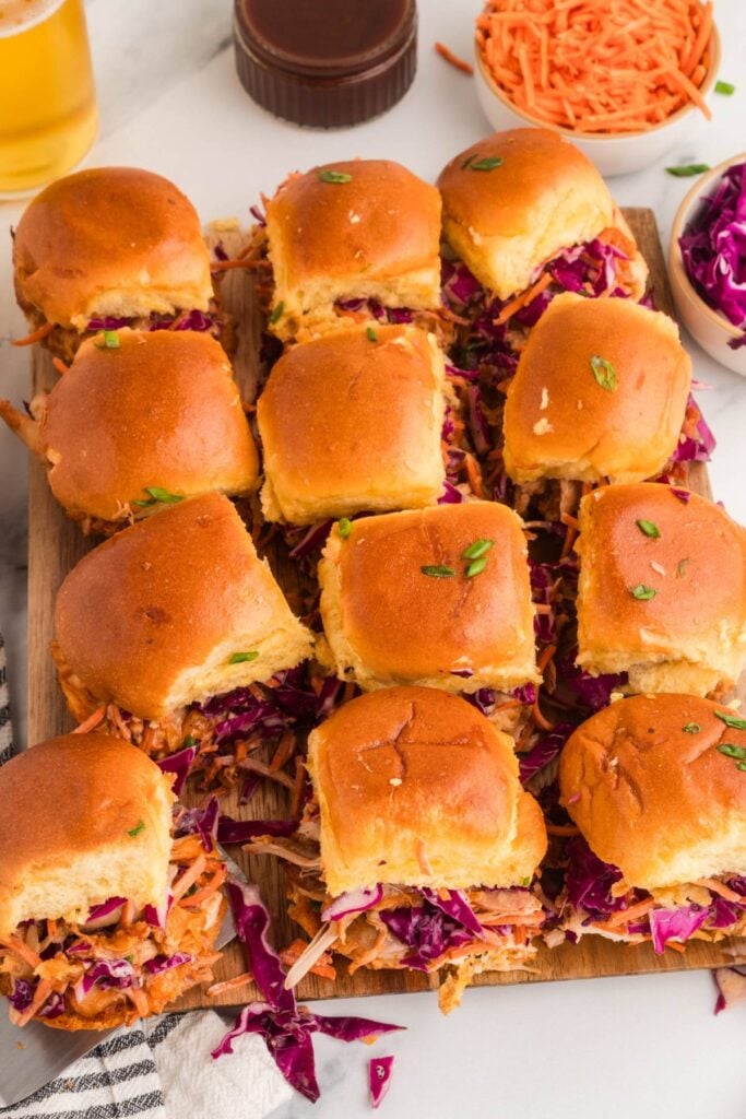 Twelve BBQ chicken sliders with coleslaw arranged on a wooden board.