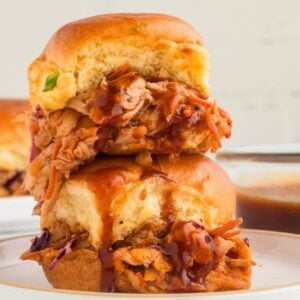 Two BBQ chicken sliders stacked on a plate with coleslaw and sauce dripping.