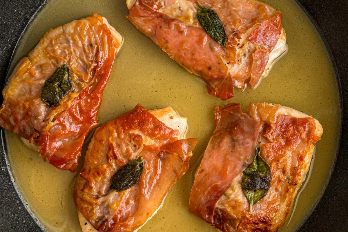 Four chicken cutlets wrapped in prosciutto and sage in a skillet with sauce.