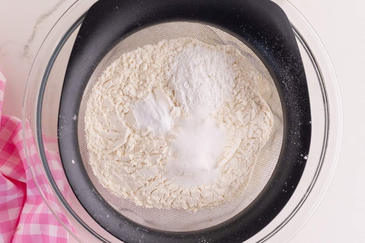 Cake flour, baking powder, salt, and baking soda in a mesh strainer over a bowl.