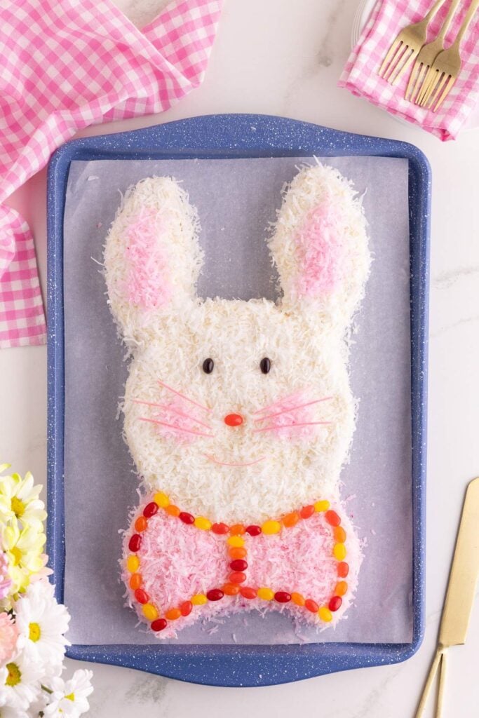 Finished coconut bunny cake with pink ears and jellybean bowtie on a blue tray.