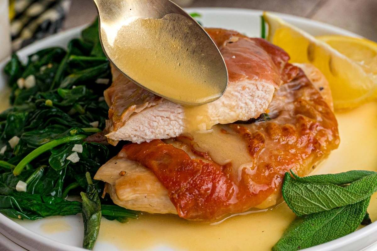 Spoon drizzling butter sauce over sliced chicken saltimbocca with sautéed spinach.