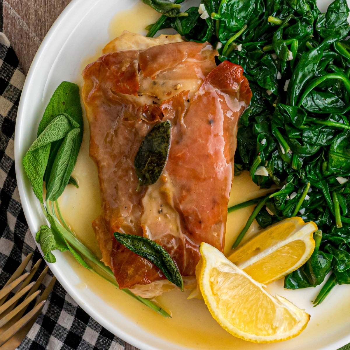 Single chicken saltimbocca on a plate with spinach and lemon wedges.