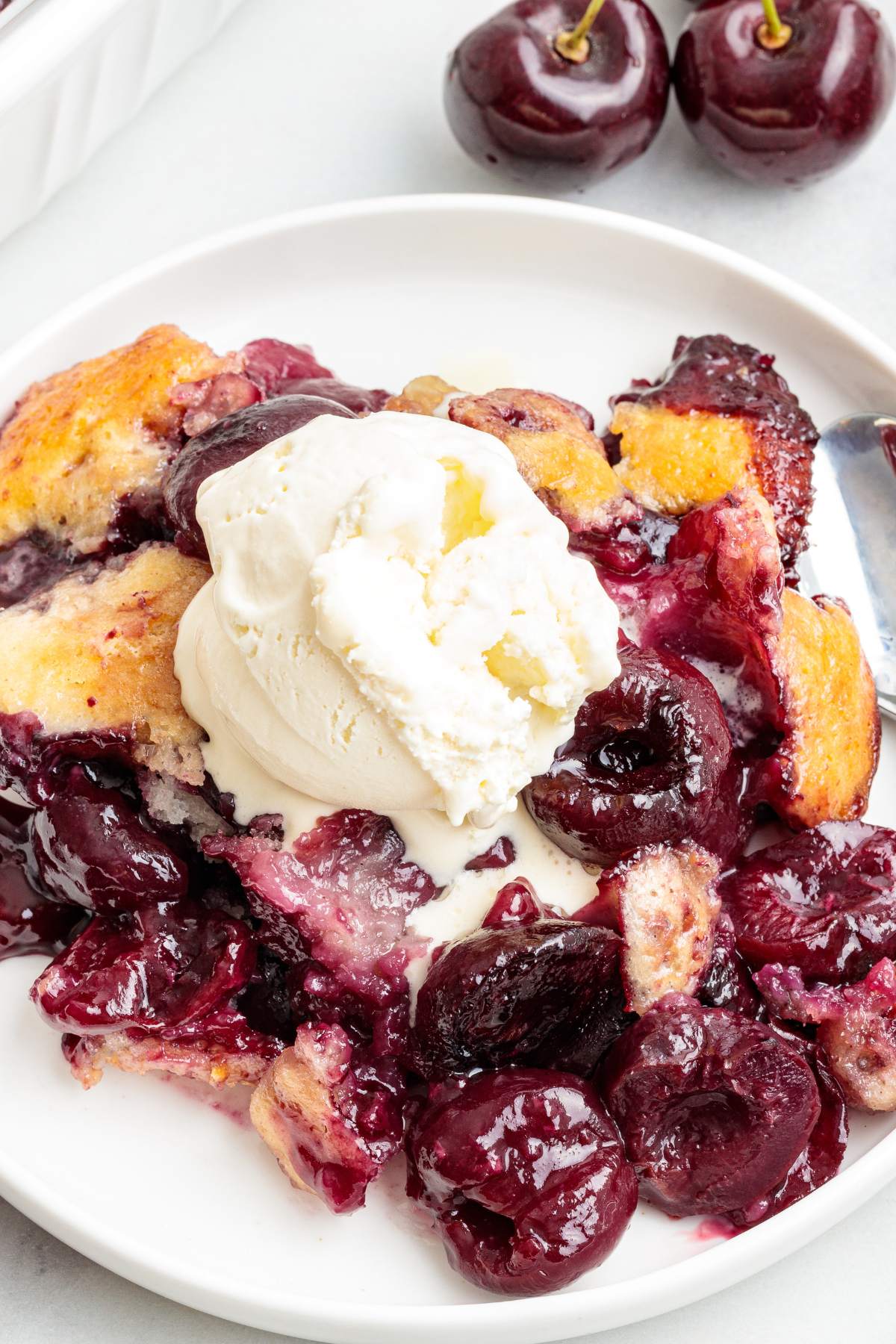 Baked cherry cobbler topped with vanilla ice cream on a white plate.