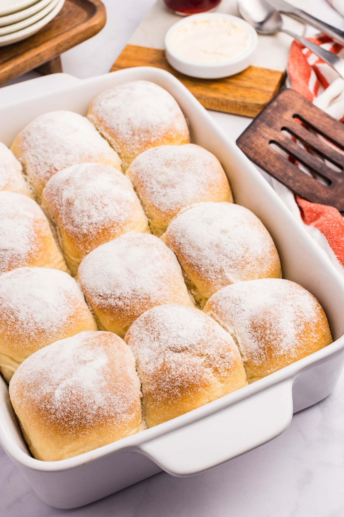 A pan of twelve freshly baked snowflake rolls dusted with flour in a white ceramic baking dish, with a small bowl of butter and a wooden spatula in the background.