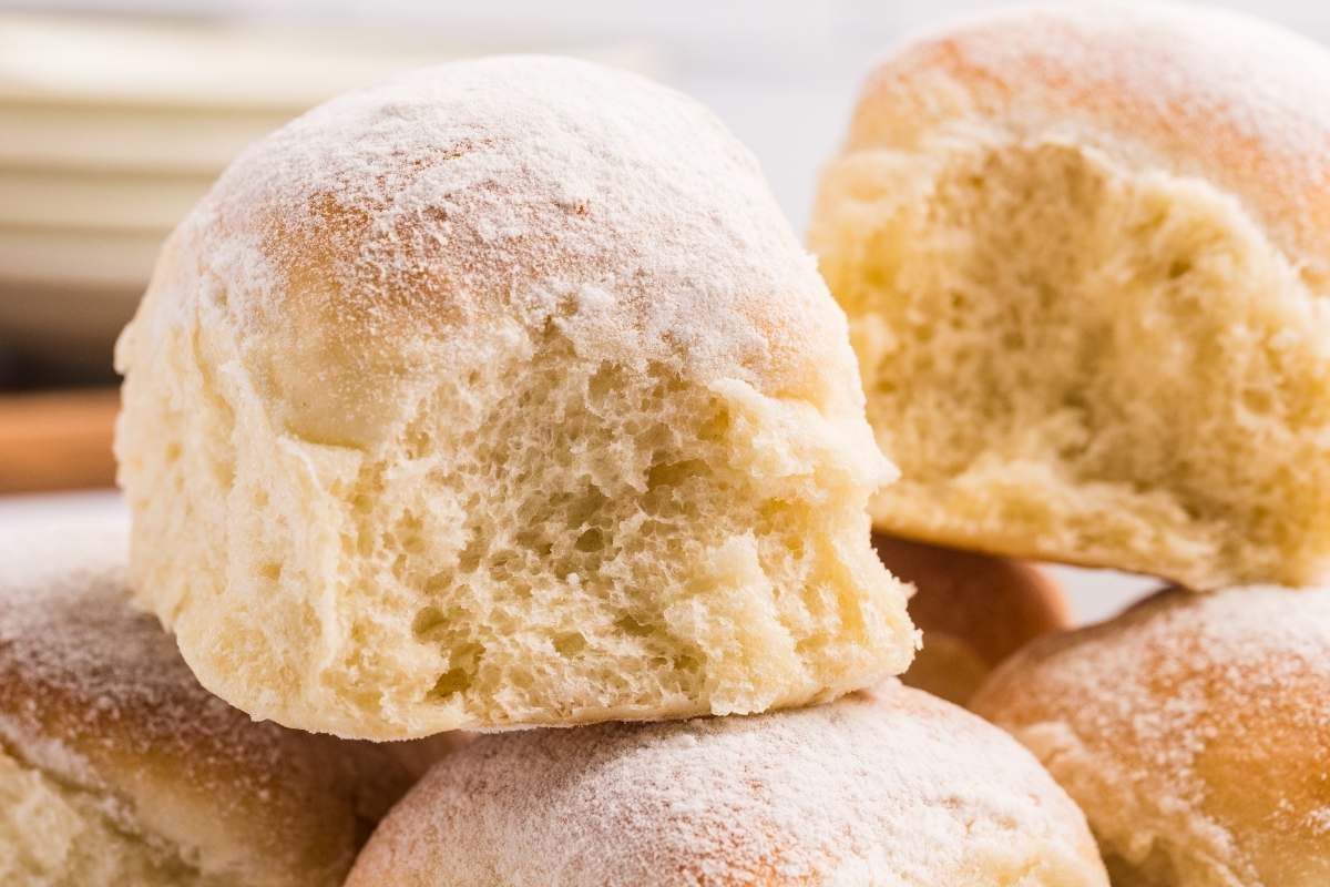 A split snowflake roll showing a soft, fluffy interior, stacked on top of more flour-dusted rolls on a wooden surface.
