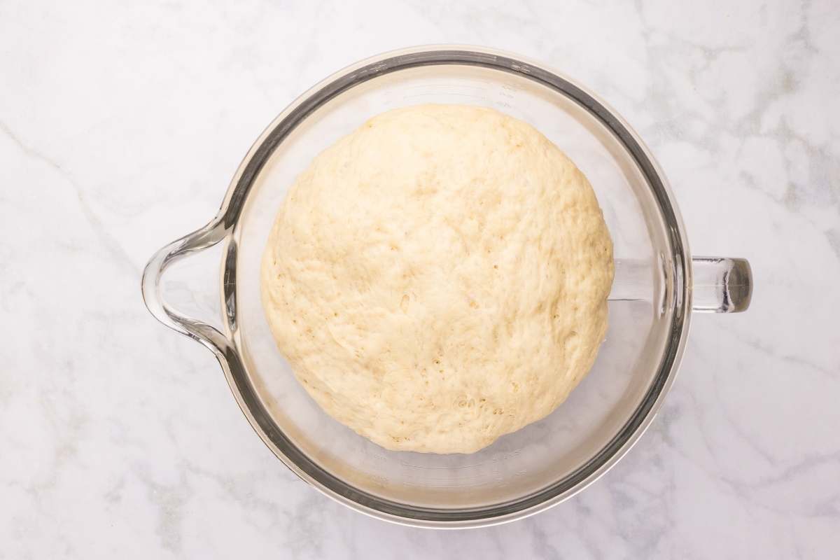 Overhead view of snowflake roll dough that has doubled in size in a glass stand mixer bowl on a marble surface.