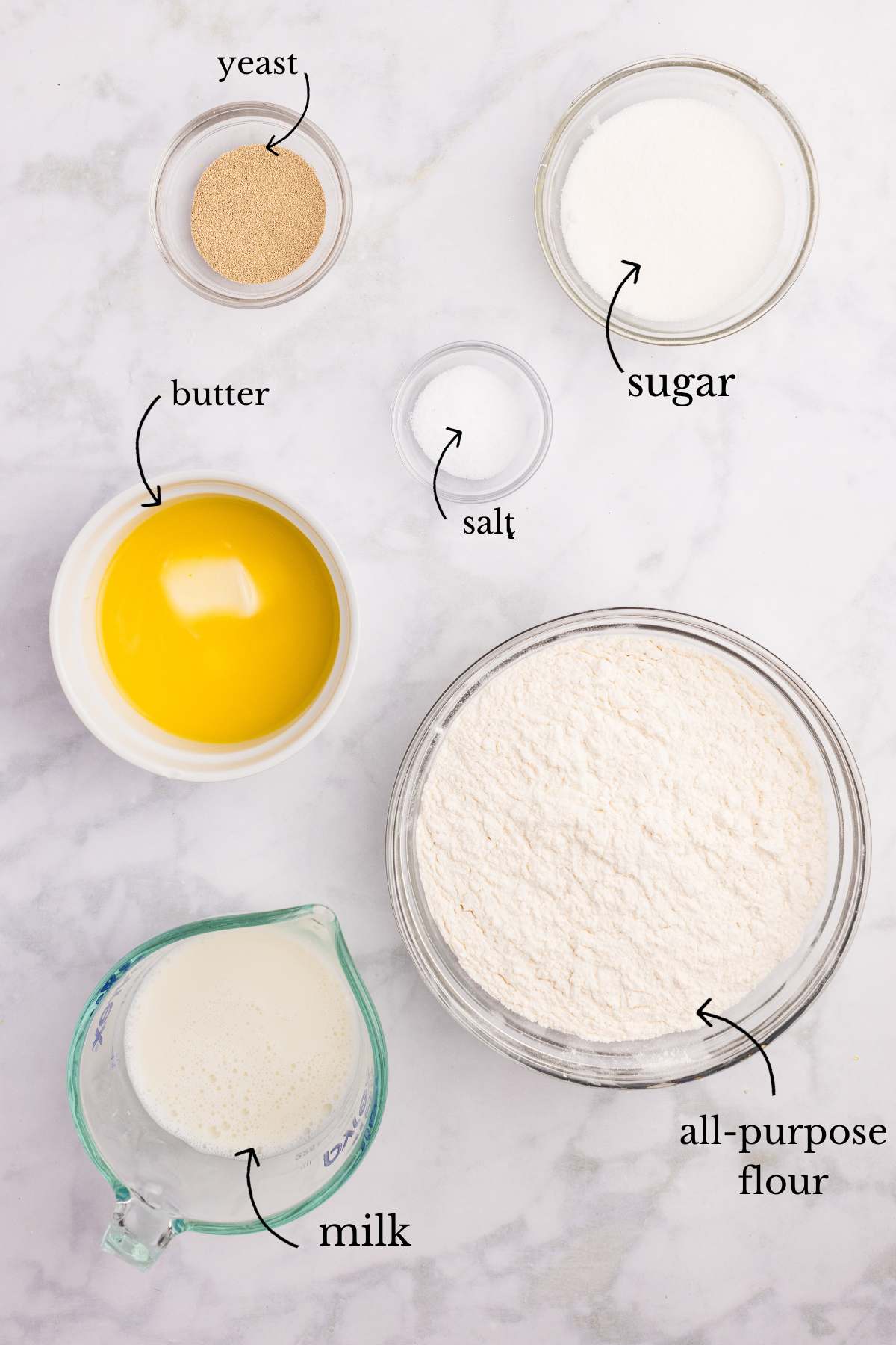 
Overhead flat lay of snowflake roll ingredients in glass bowls on a marble surface, labeled with arrows: yeast, sugar, butter, salt, all-purpose flour, and milk.