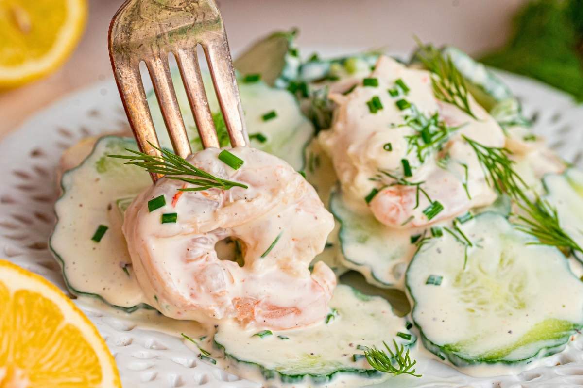 Fork lifting a creamy coated shrimp from a white plate of cucumber salad.
