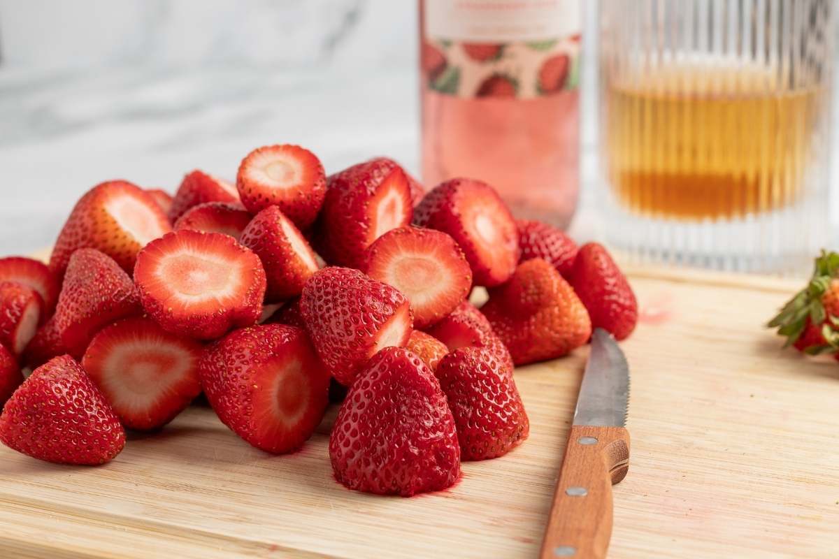 Hulled strawberries on a cutting board with a knife and two bottles.