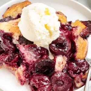 Baked cherry cobbler topped with vanilla ice cream on a white plate.