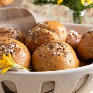Close-up of rye dinner rolls in a white ceramic basket, topped with caraway seeds and flaky salt.