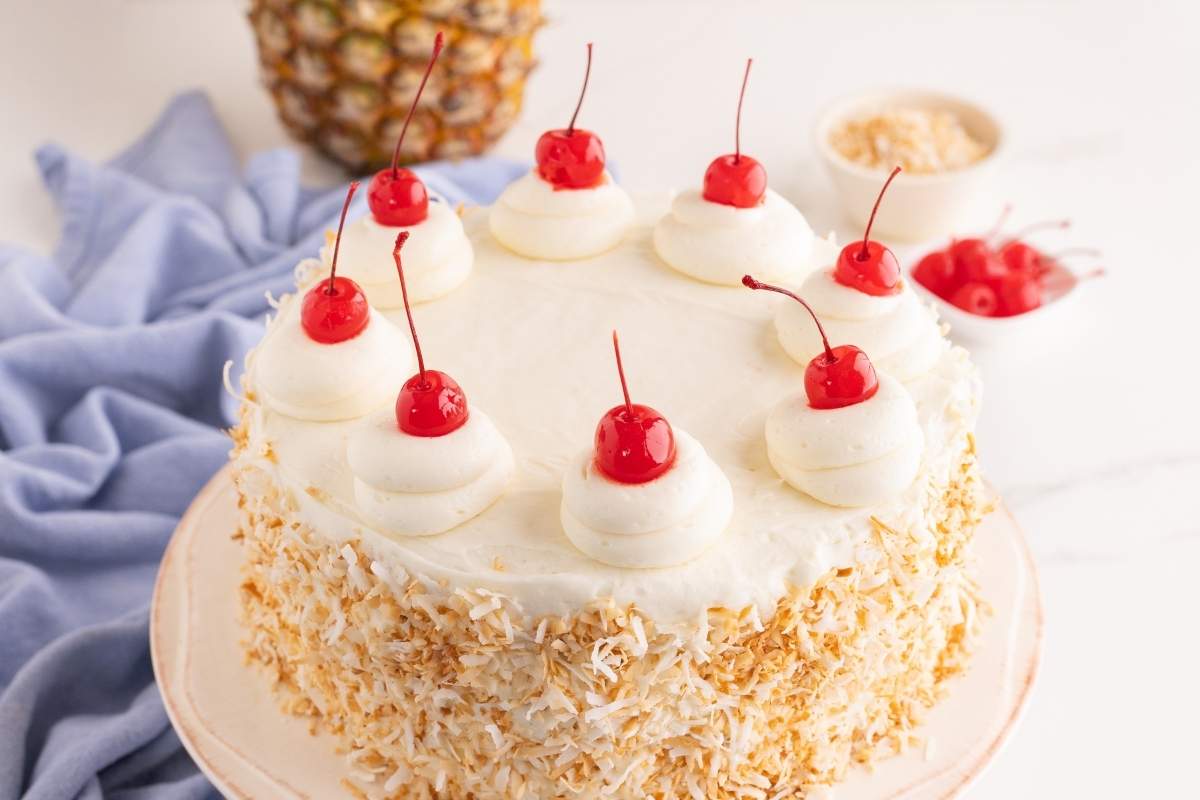Piña colada layer cake coated with toasted coconut and topped with whipped cream swirls and maraschino cherries.