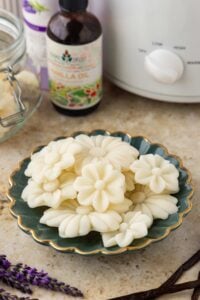 White flower-shaped wax melts arranged on a green scalloped dish beside vanilla oil, lavender sprigs, and a slow cooker.