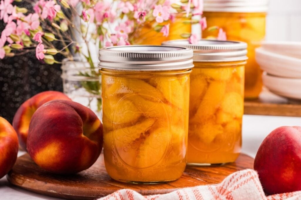 Mason jars filled with sliced peaches in syrup sitting on a red striped towel beside fresh peaches.