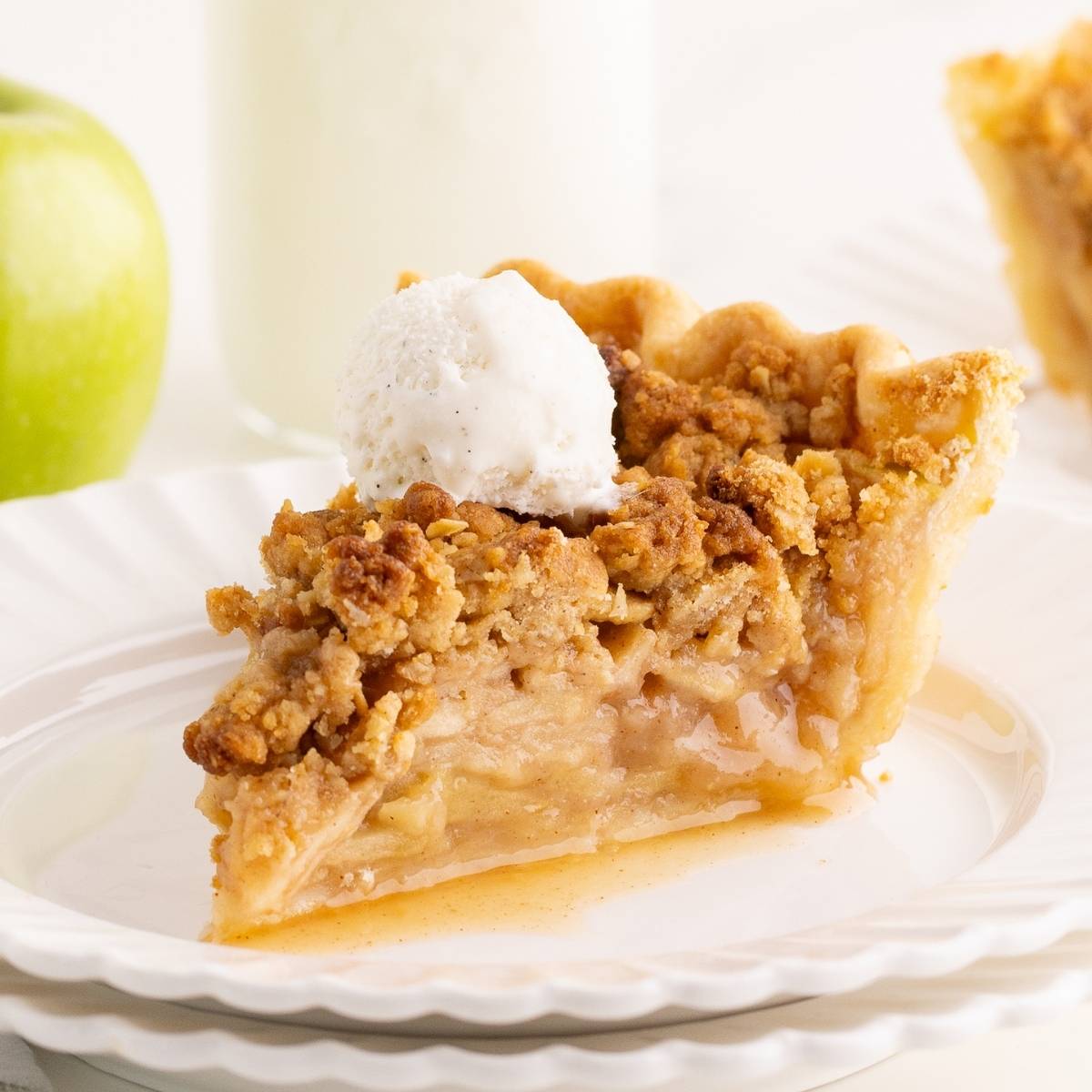 Slice of Dutch apple pie with golden crumble topping and a scoop of vanilla ice cream on a white plate.
