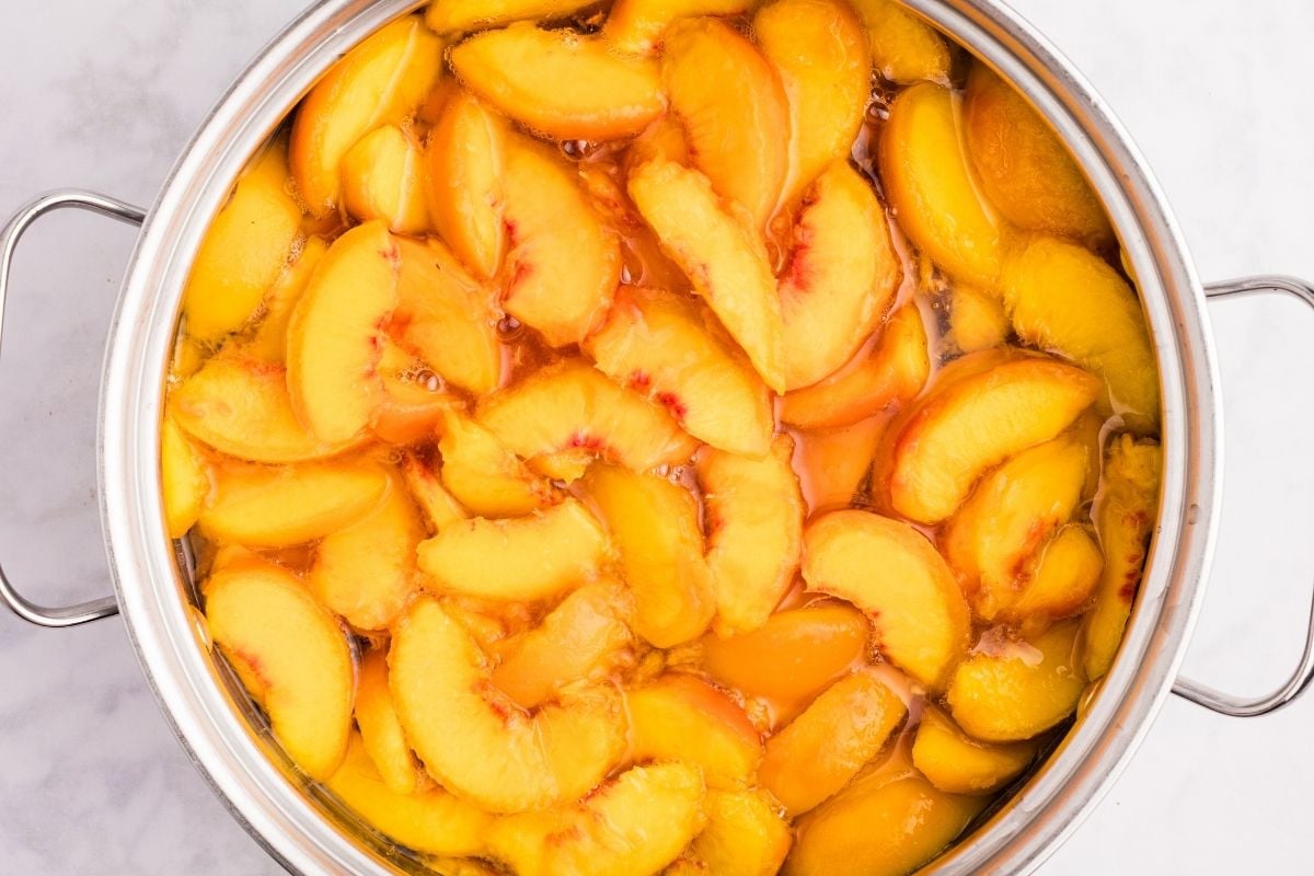 Sliced peaches simmering in golden syrup inside a large stainless steel pot on a marble surface.