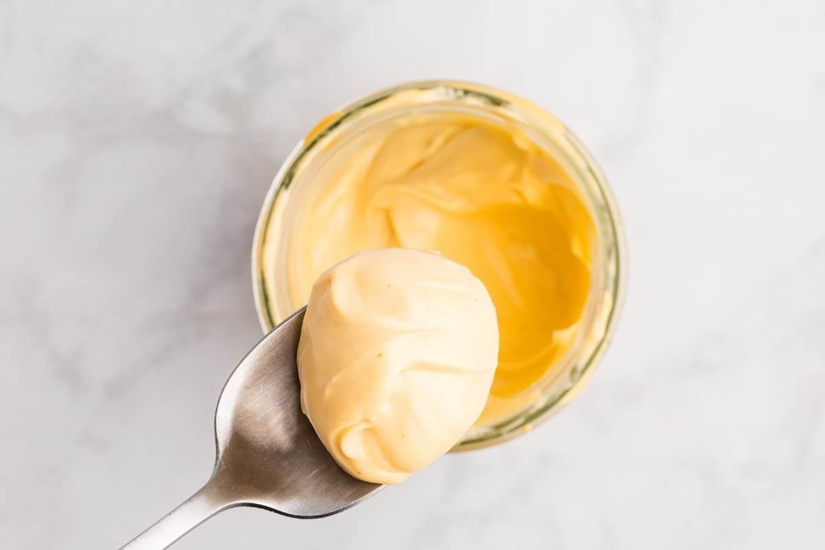A spoon lifting a dollop of pale yellow homemade mayonnaise from a glass jar for tasting.