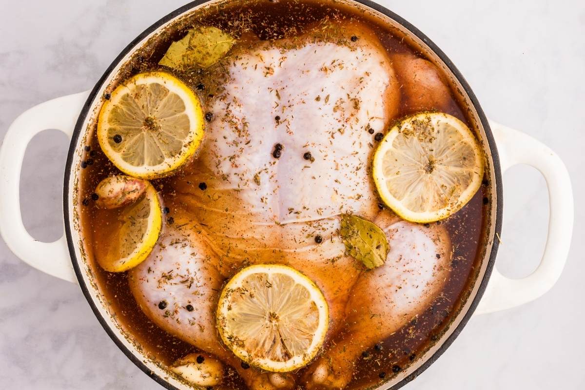 Chicken brining in a white pot with lemon slices, bay leaves, peppercorns, and herbs in amber liquid.