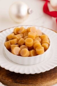 Round golden butterscotch candies piled in a white fluted dish on a wooden board with Christmas ornaments behind.