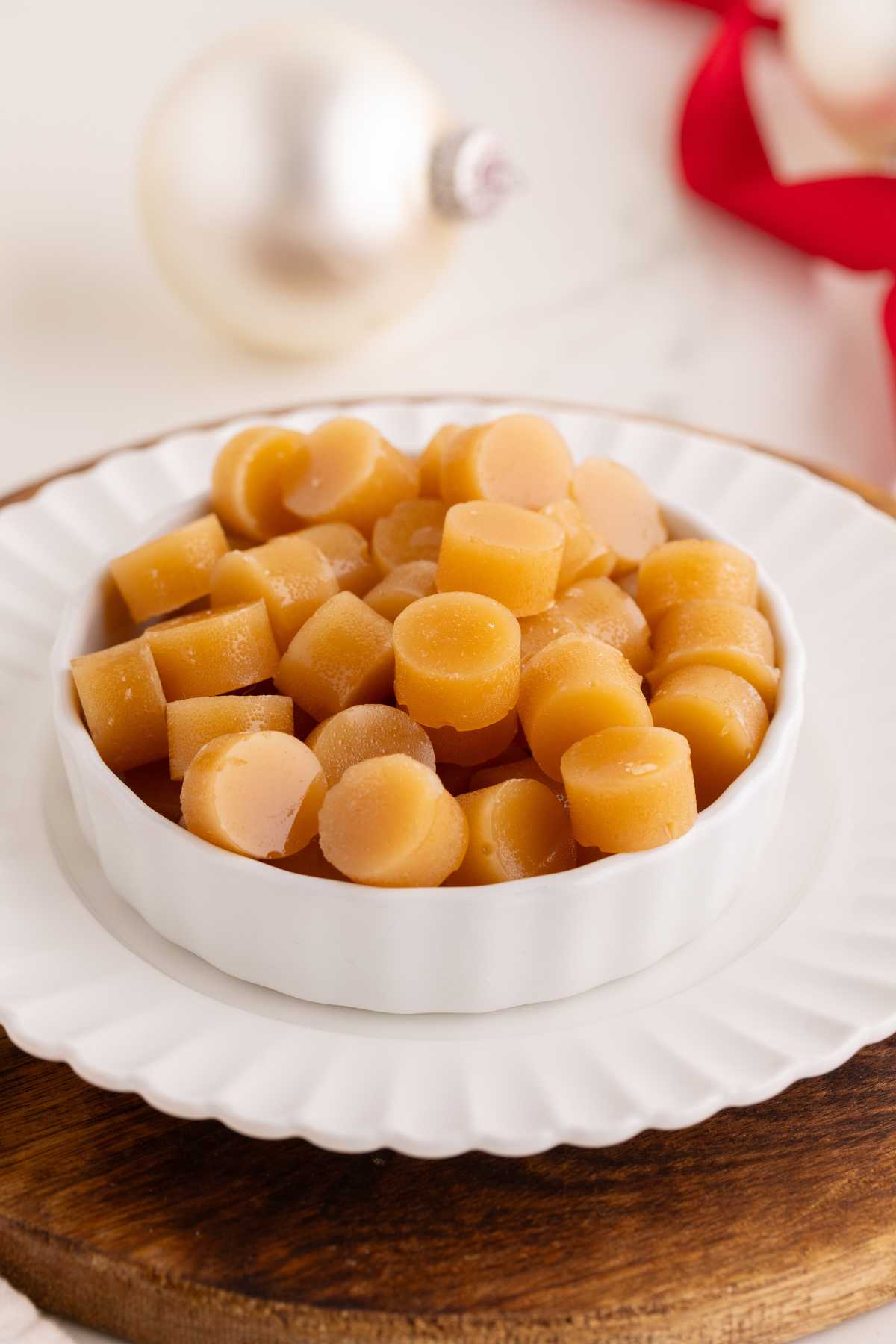 Round golden butterscotch candies piled in a white fluted dish on a wooden board with Christmas ornaments behind.