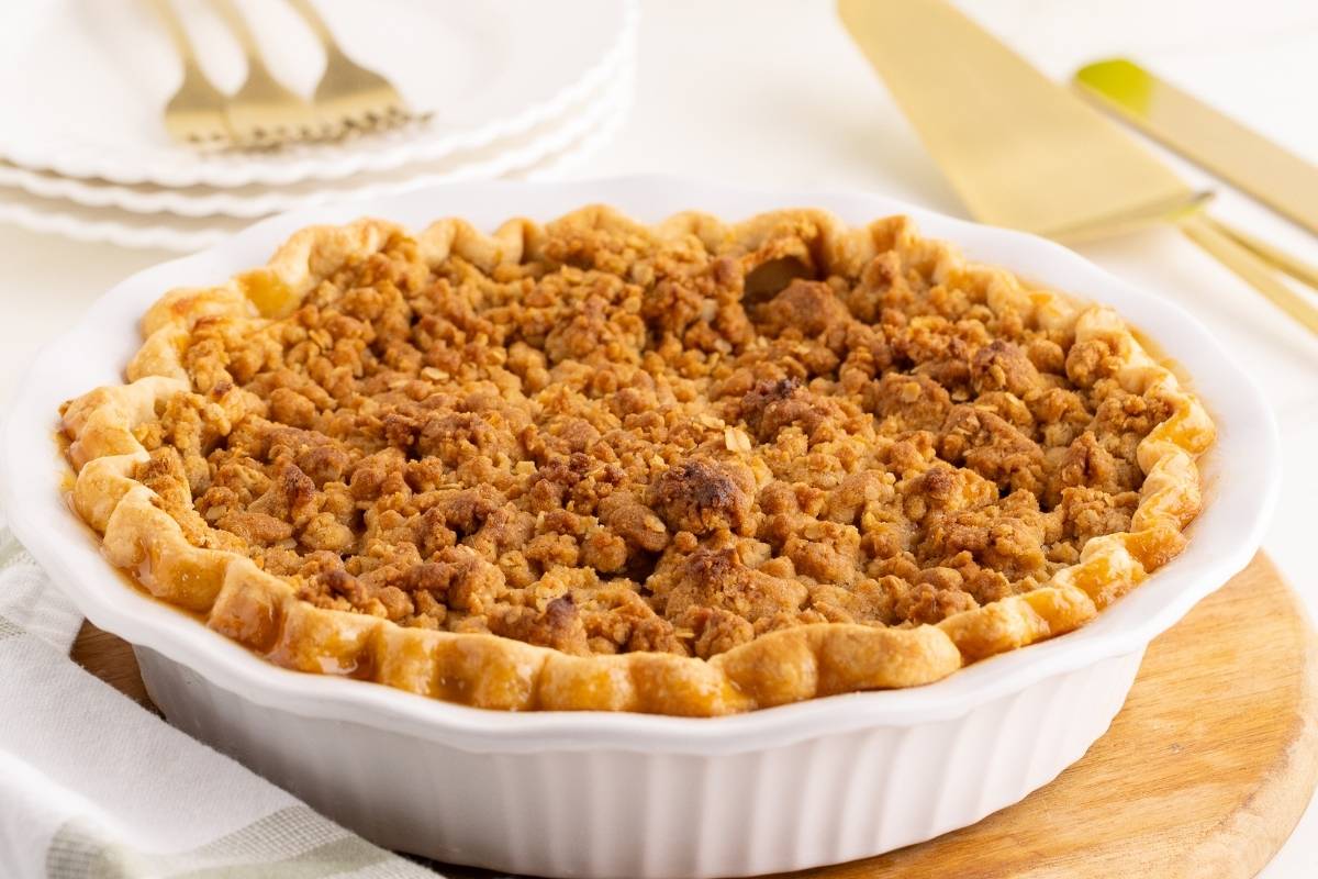 Golden crumb-topped apple pie cooling in a white fluted dish on a wooden board.