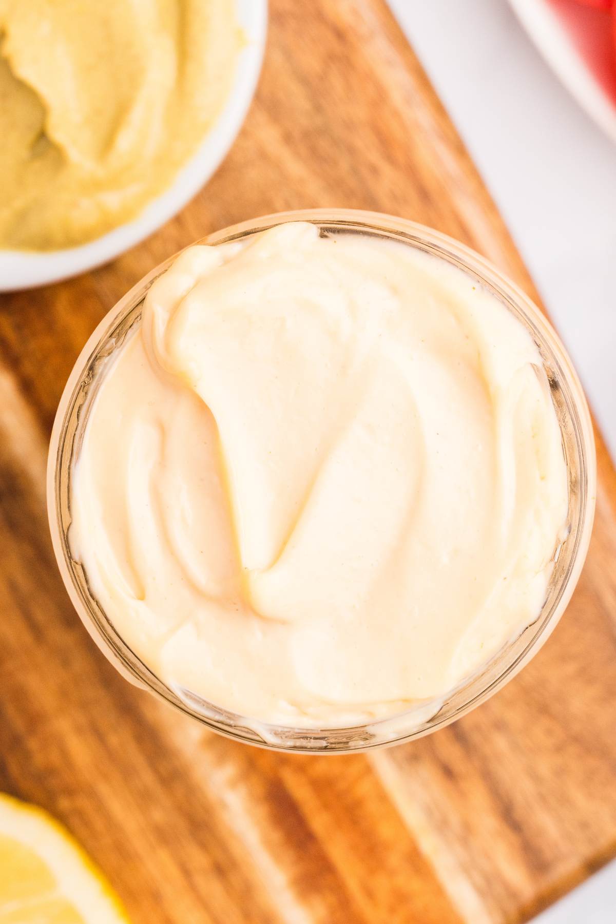 Creamy homemade mayonnaise in a glass jar on a wooden board beside mustard and lemon.