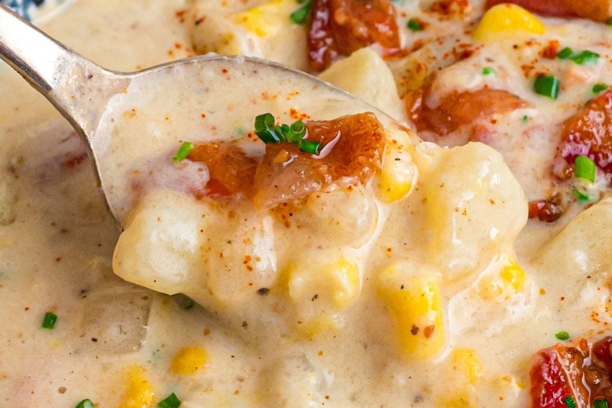 Spoon lifting creamy corn chowder with chunks of potato, bacon, and chives for tasting.
