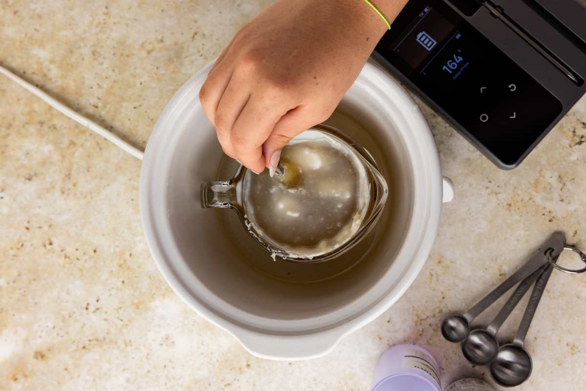 Hand stirring melted beeswax in a glass measuring cup inside a white sous vide bath at 164 degrees.