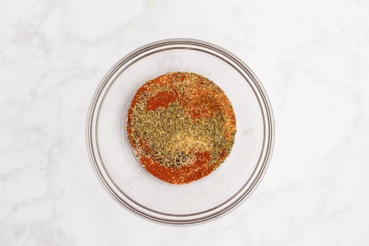 Paprika, dried herbs, garlic powder, and other seasonings layered in a glass bowl before mixing.