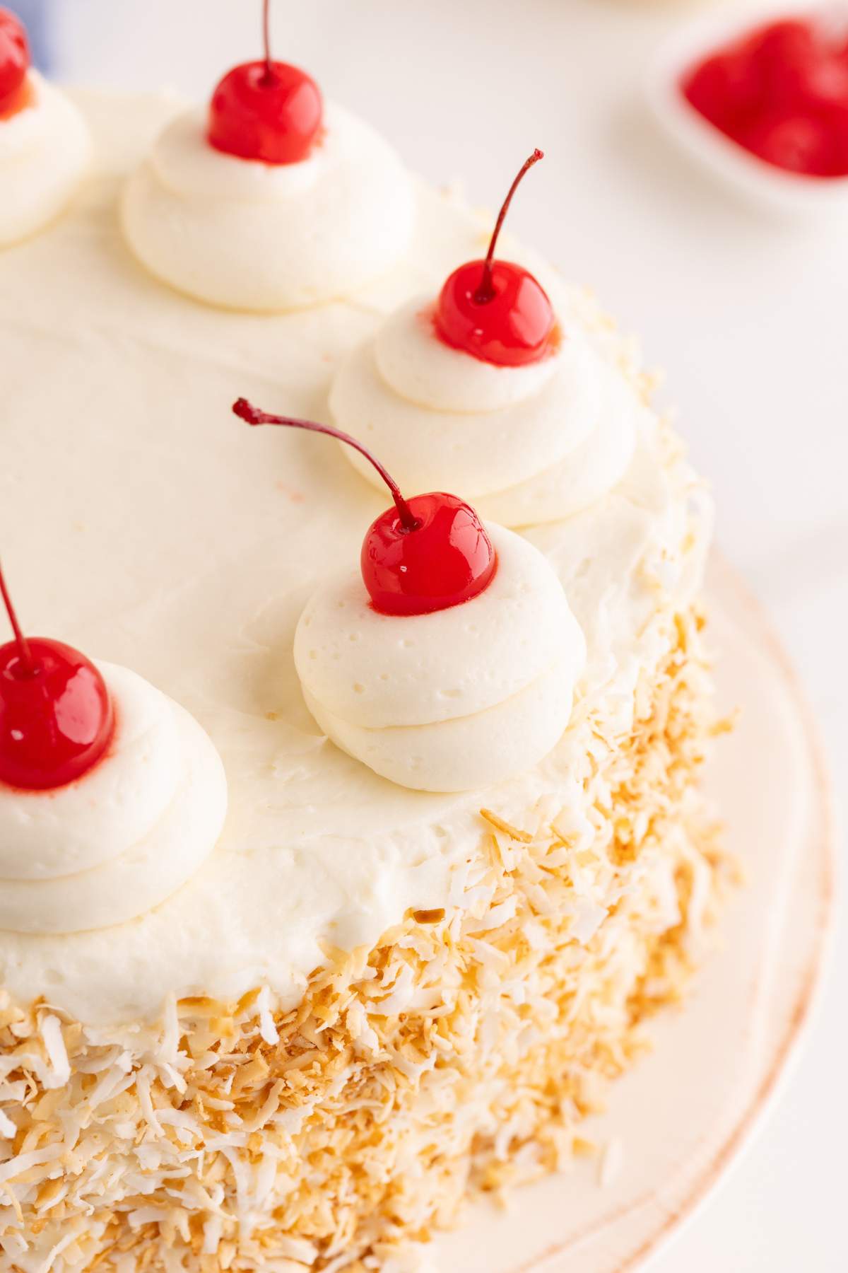 White-frosted layer cake topped with maraschino cherries on piped buttercream swirls and toasted coconut around the edges.
