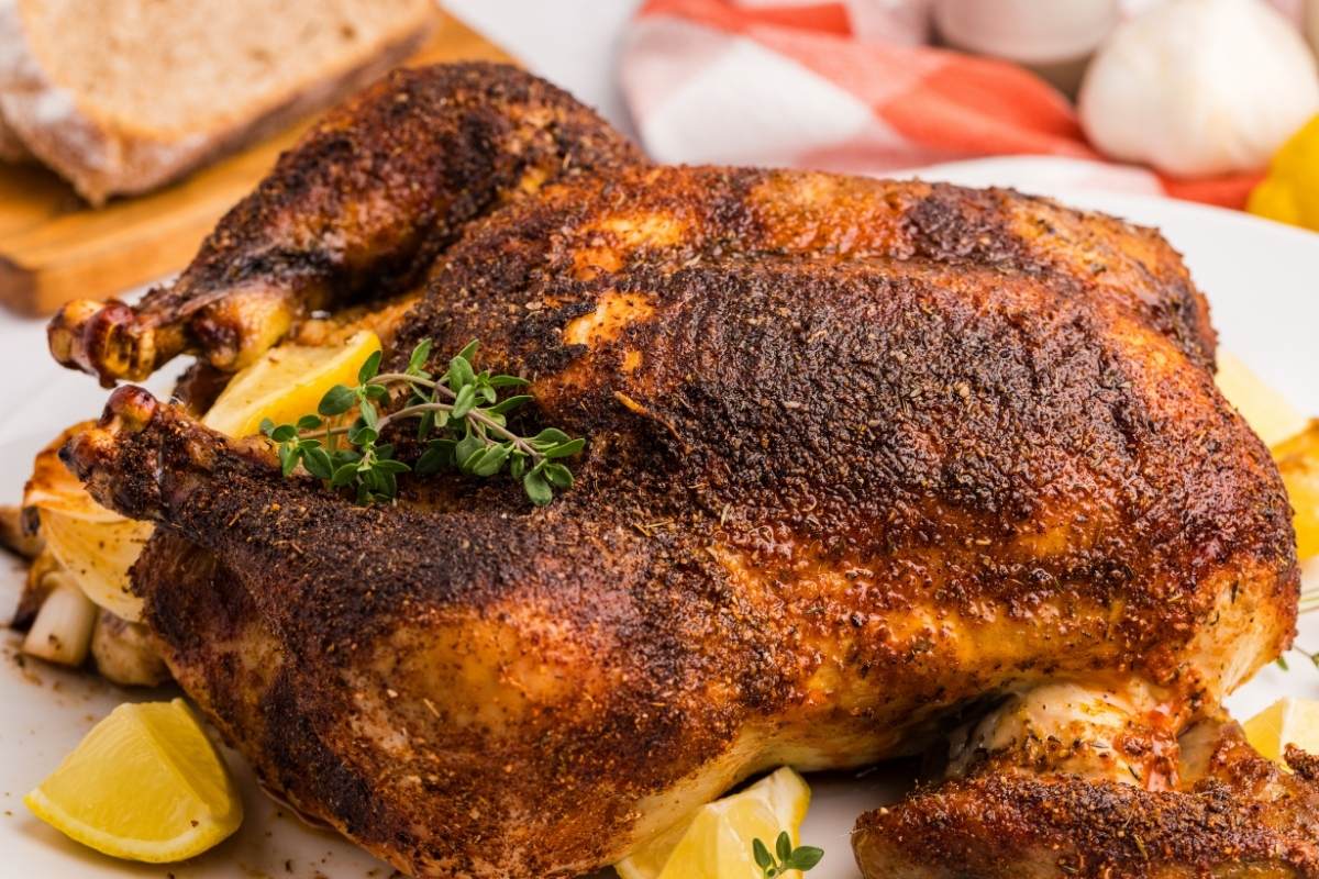 Whole spice-rubbed rotisserie chicken resting on a platter with lemon wedges and fresh thyme sprigs.