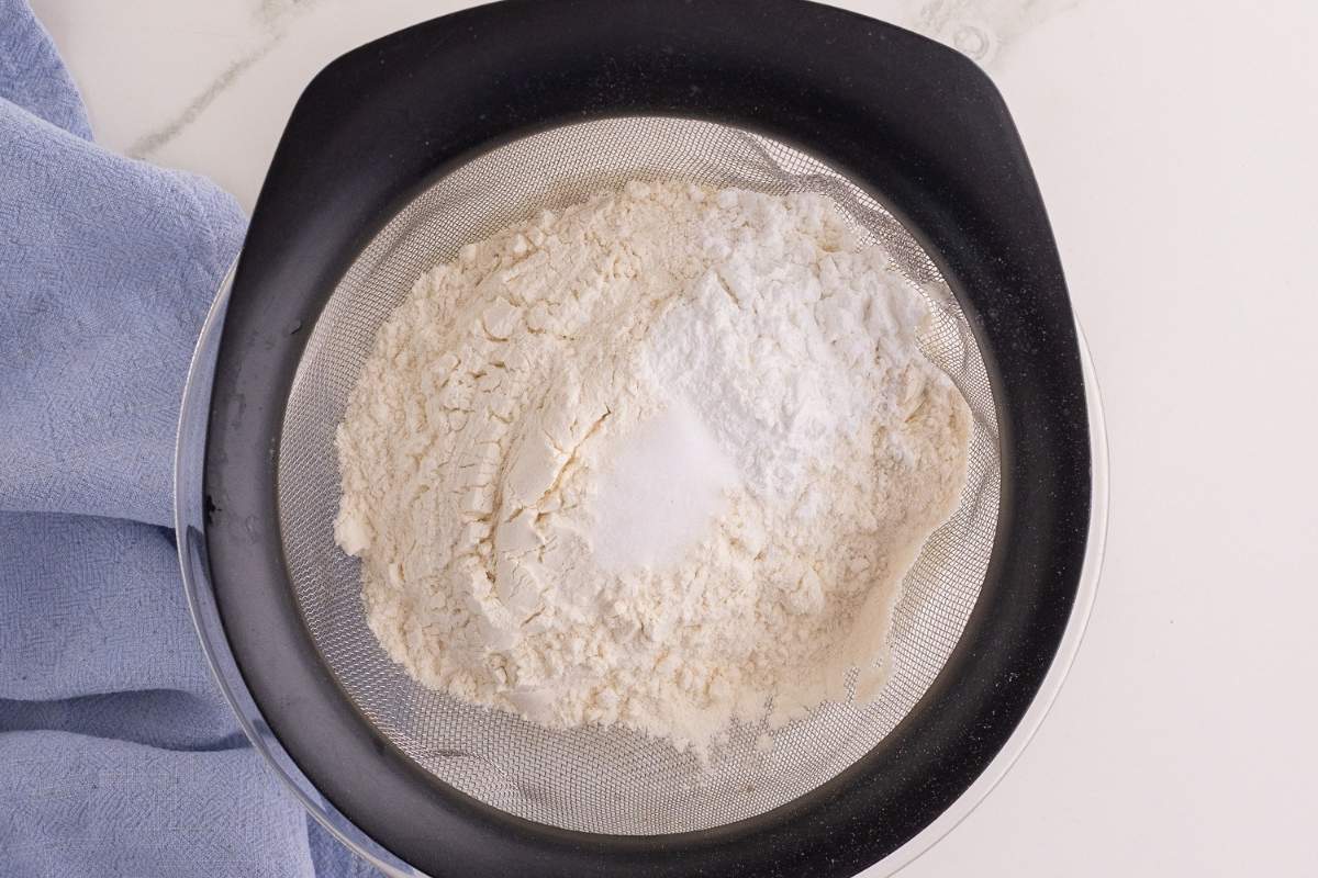 Flour and leavening being sifted through a mesh strainer over a black bowl beside a blue towel.