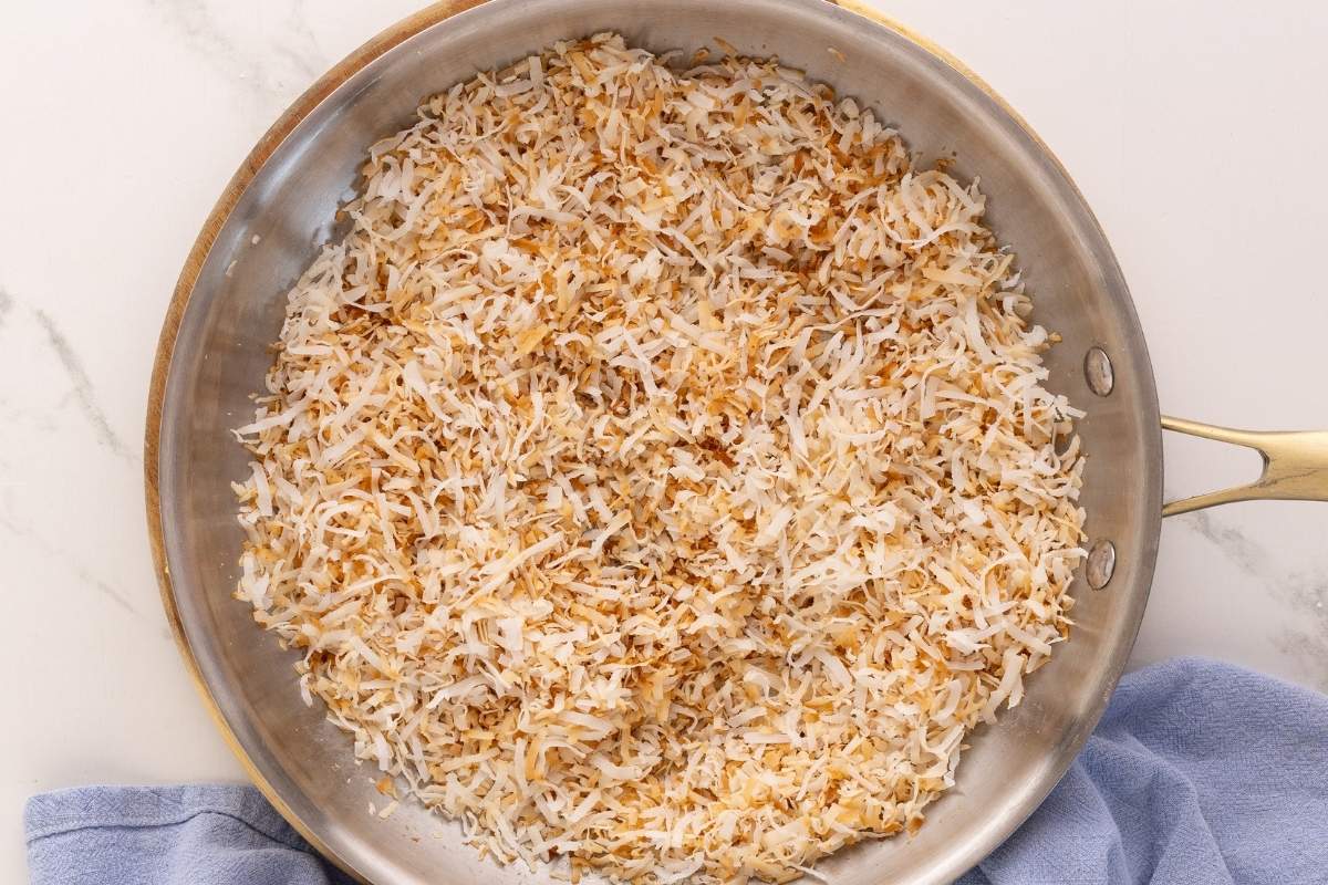 Shredded coconut toasting to golden brown in a stainless steel skillet on a marble surface.
