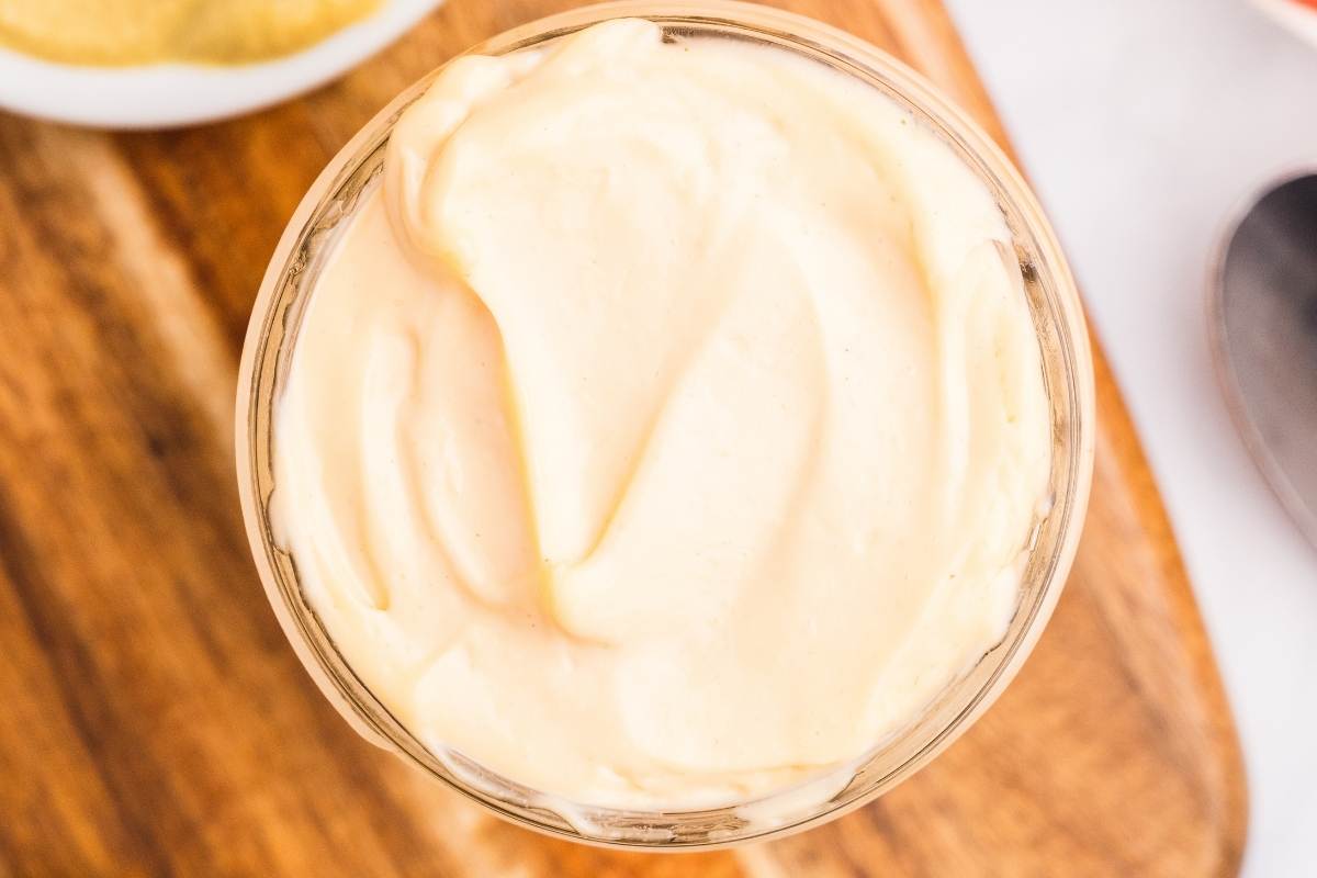 Overhead view of creamy pale yellow homemade mayonnaise swirled in a glass jar on a wooden board.