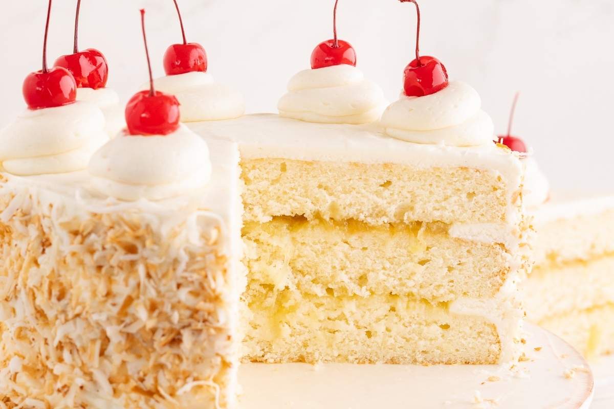 Sliced three-layer pina colada cake showing pineapple filling, toasted coconut sides, and buttercream swirls topped with maraschino cherries.
