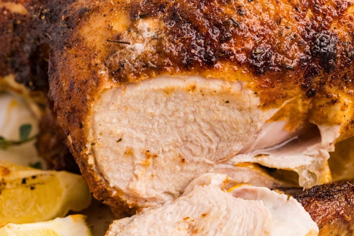 Close-up of sliced rotisserie-style chicken with crispy seasoned golden-brown skin and juicy white meat beside roasted lemon wedges.