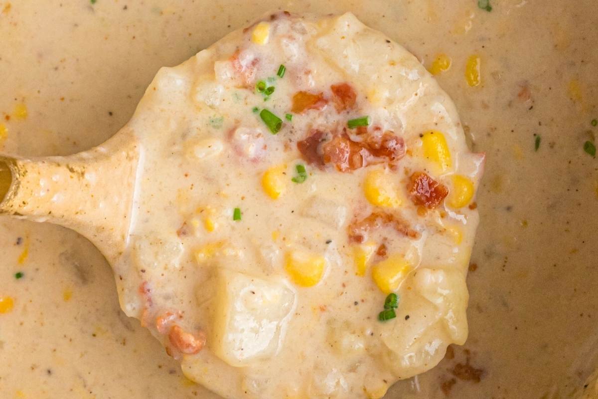 Ladle scooping creamy corn chowder with bacon bits, potato chunks, and fresh chives from a pot.