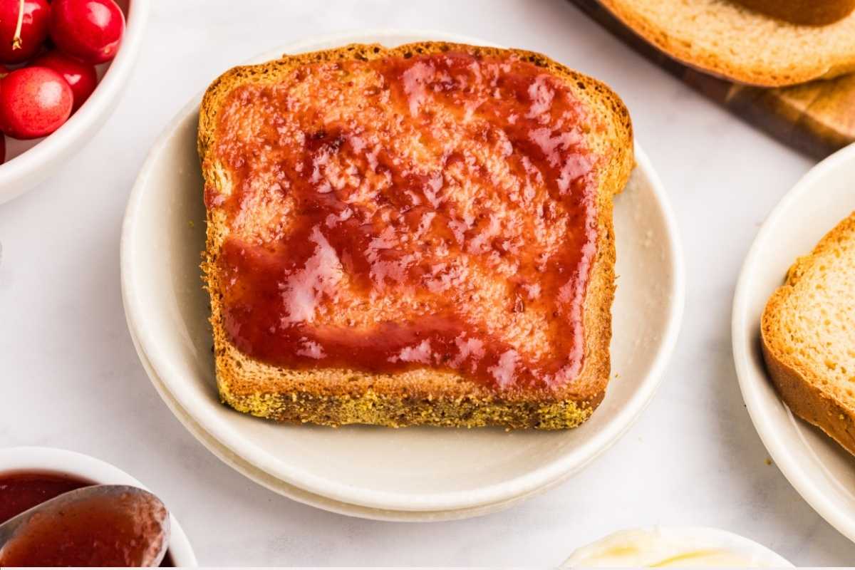 Slice of toasted English muffin bread topped with strawberry jam on a white plate beside cherries.