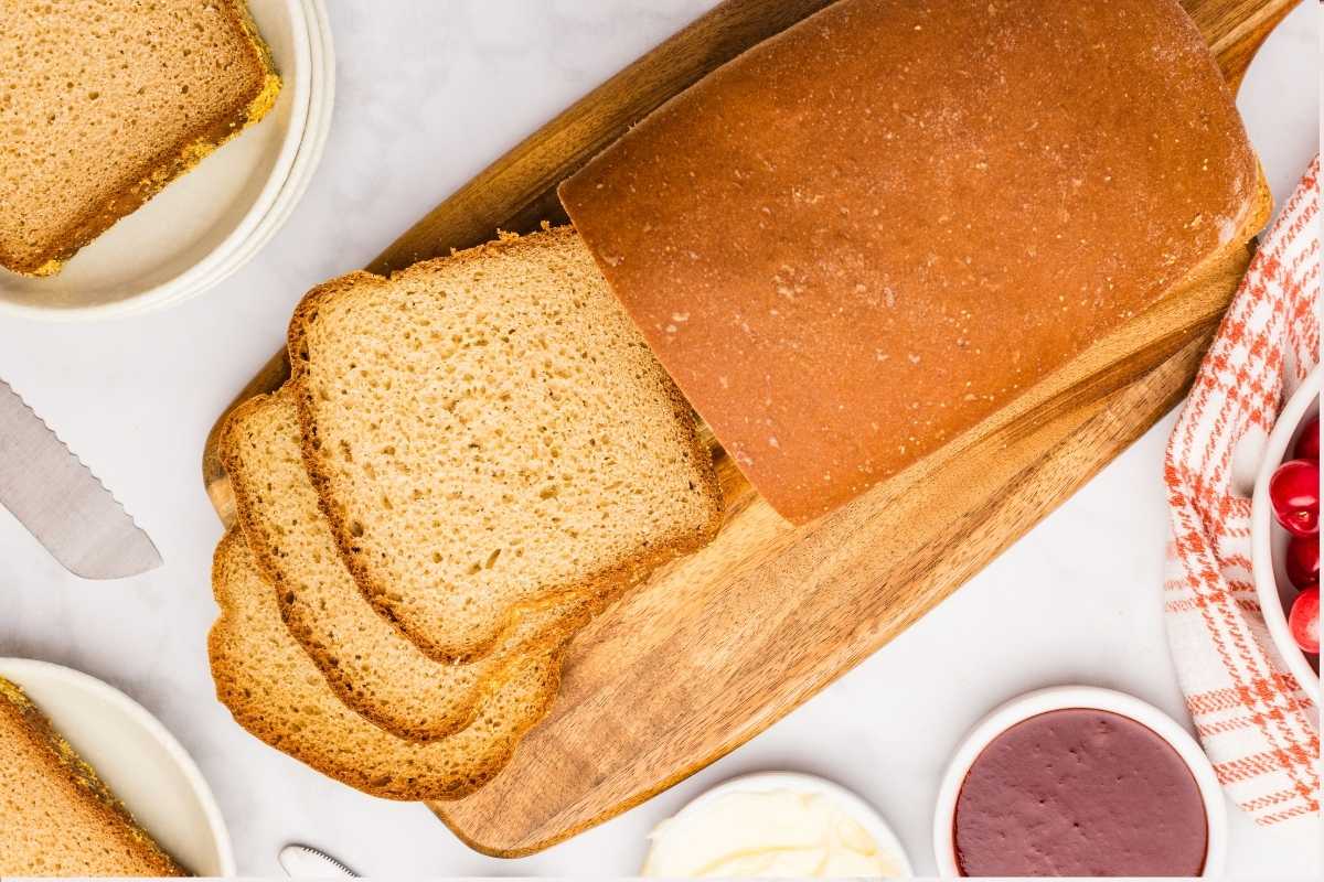 Sliced golden loaf of English muffin bread on a wooden board with butter, jam, and cranberries nearby.
