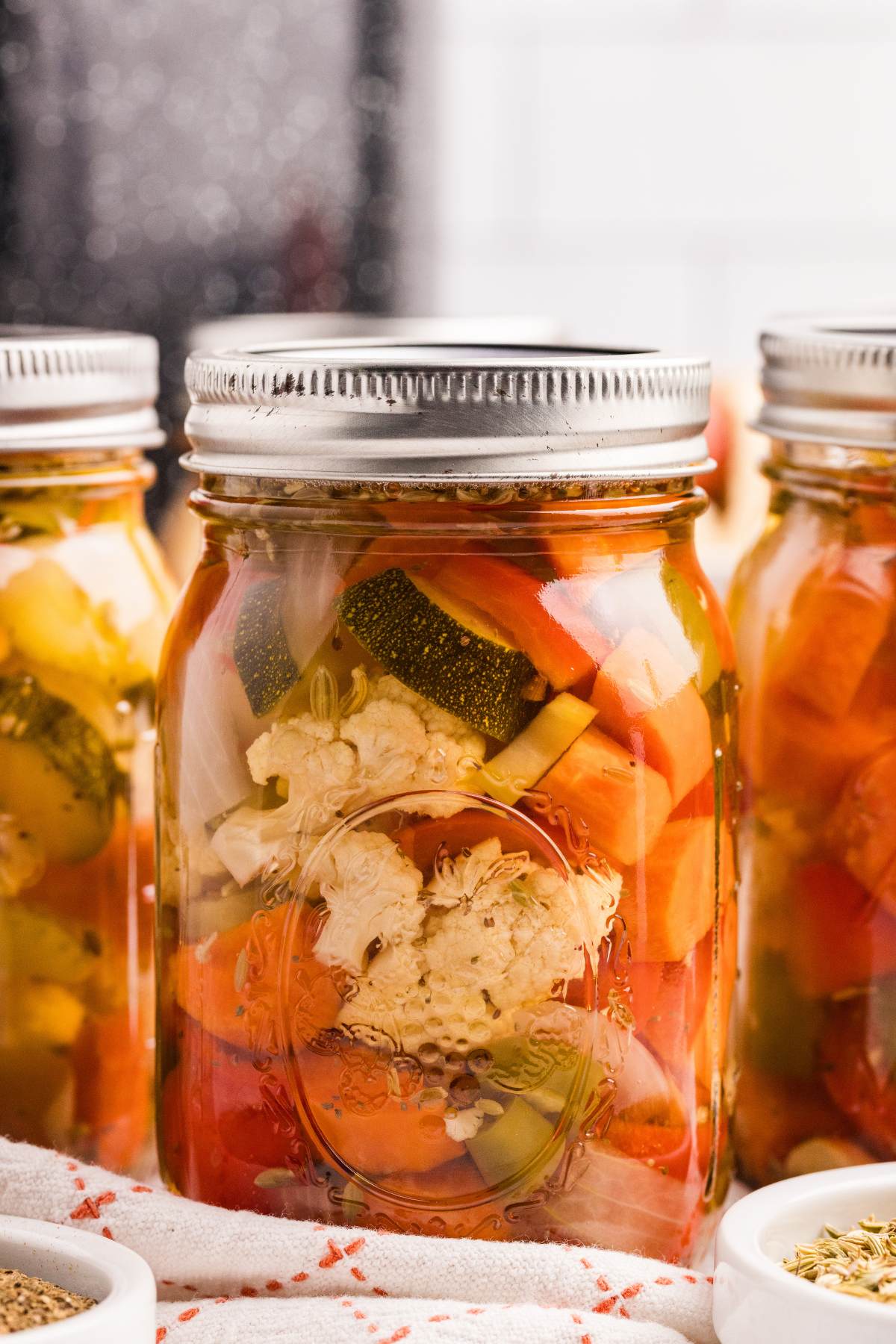 Mason jars filled with colorful pickled cauliflower, carrots, and peppers beside fennel seeds and bread.
