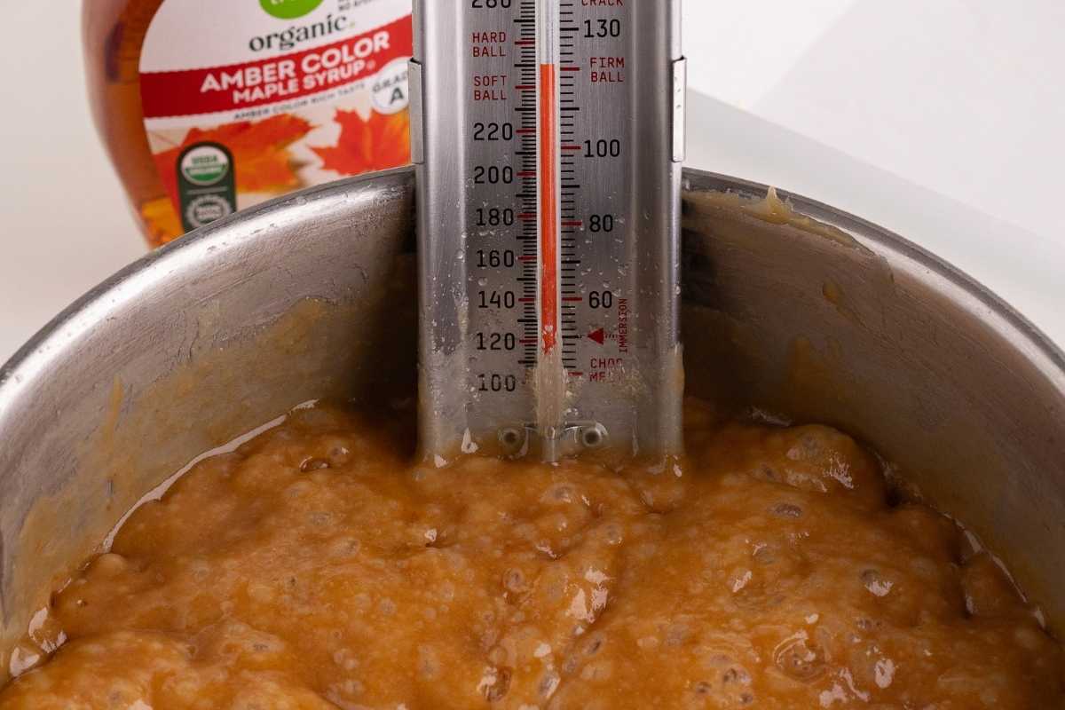 Candy thermometer clipped inside a saucepan of bubbling caramel mixture with amber maple syrup bottle behind.
