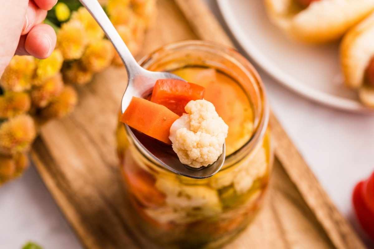 Spoon lifting pickled cauliflower and carrot from a glass jar of giardiniera on a wooden board.