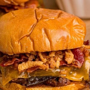 Close-up of a BBQ beef sandwich with crispy fried onions, bacon, melted cheese, and shredded beef on a glossy brioche bun.