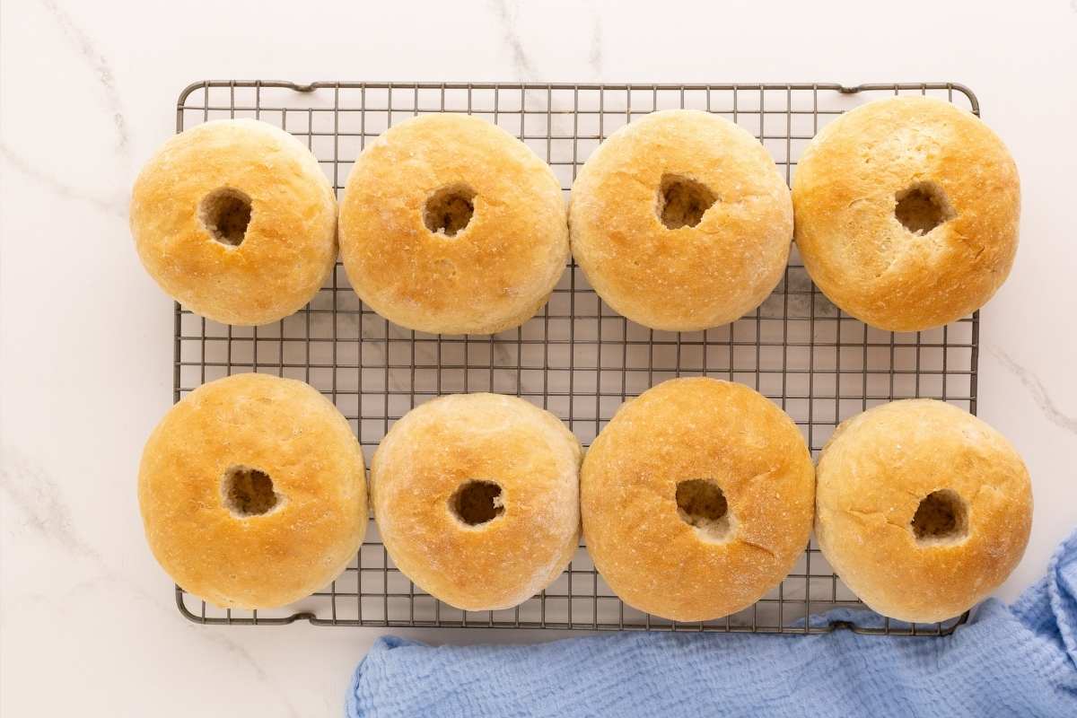 Eight golden baked buns with cut center holes cooling on a wire rack beside a blue napkin.